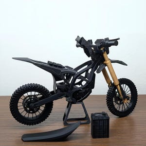 May include: A black and gold miniature motorcycle model with a detailed frame, wheels, and suspension. The model includes a seat and a separate storage box. The front forks are gold, and the tires have a textured tread.