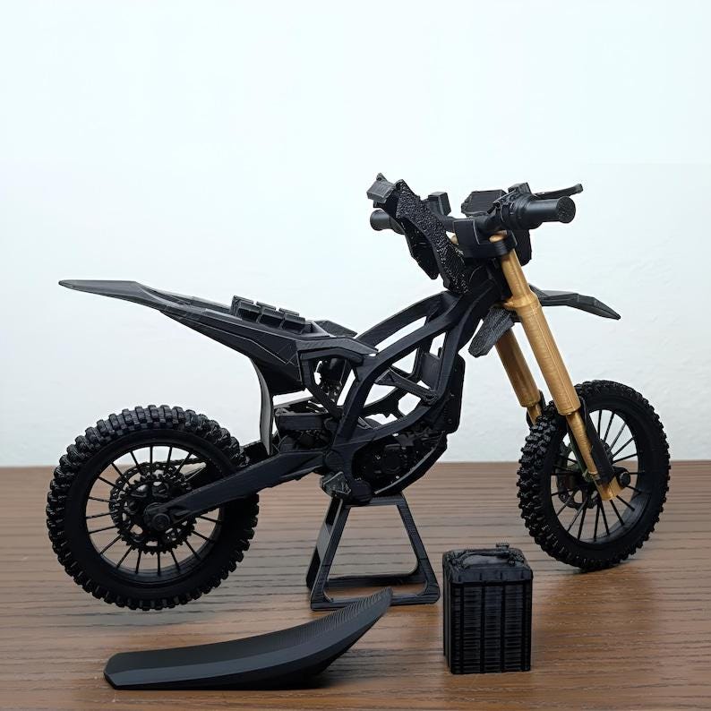 May include: A black and gold miniature motorcycle model with a detailed frame, wheels, and suspension. The model includes a seat and a separate storage box. The front forks are gold, and the tires have a textured tread.