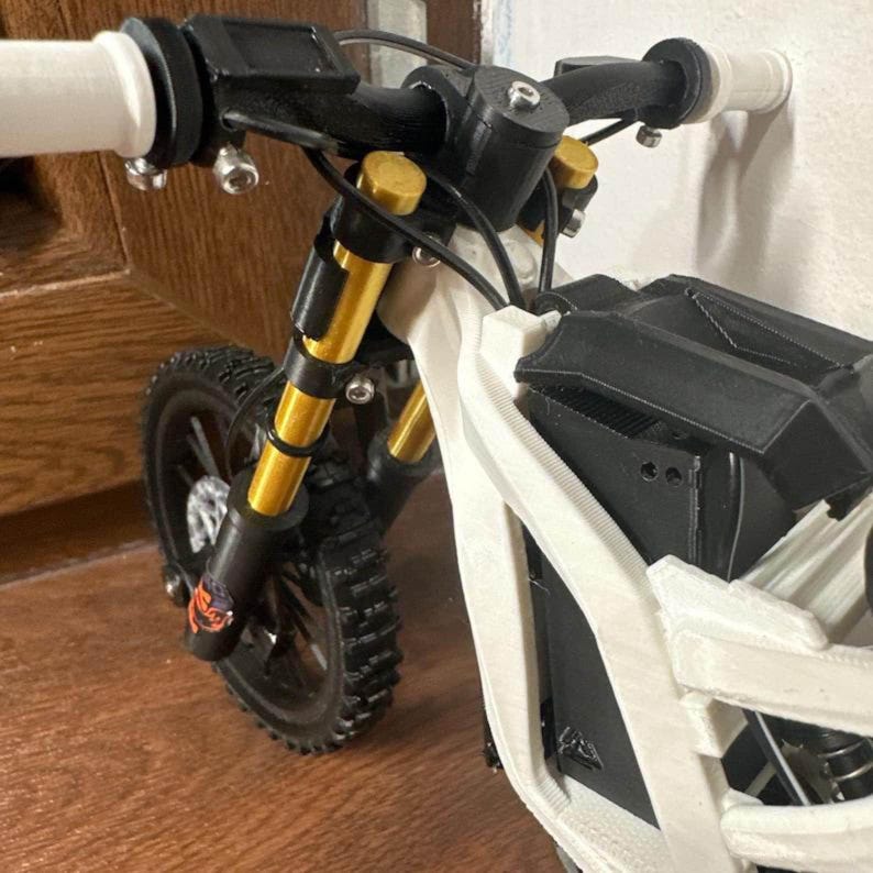 May include: A miniature black and white electric bicycle with gold and black suspension forks. The handlebars have white grips. The tires are black with a knobby tread. The frame is white and black.