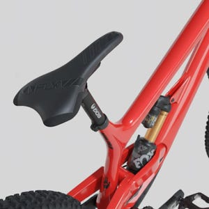 May include: Close-up of a red mountain bike frame with a black seat and suspension components. The seat is black with the brand name "FLY" visible. The suspension system includes a gold and black shock absorber.