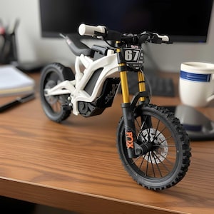 Puede incluir: Una moto de cross de juguete blanca y negra con horquillas de suspensión doradas y neumáticos negros. La moto tiene el número 67 en la parte delantera y la palabra "FOX" en naranja. El manillar y el asiento son negros.