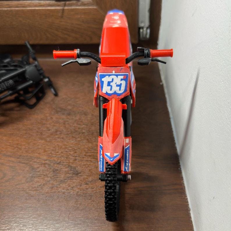 Puede incluir: Una motocicleta de juguete roja y negra con el n&uacute;mero 135 en la parte delantera. La motocicleta tiene un neum&aacute;tico negro y manillar rojo. Es un juguete.