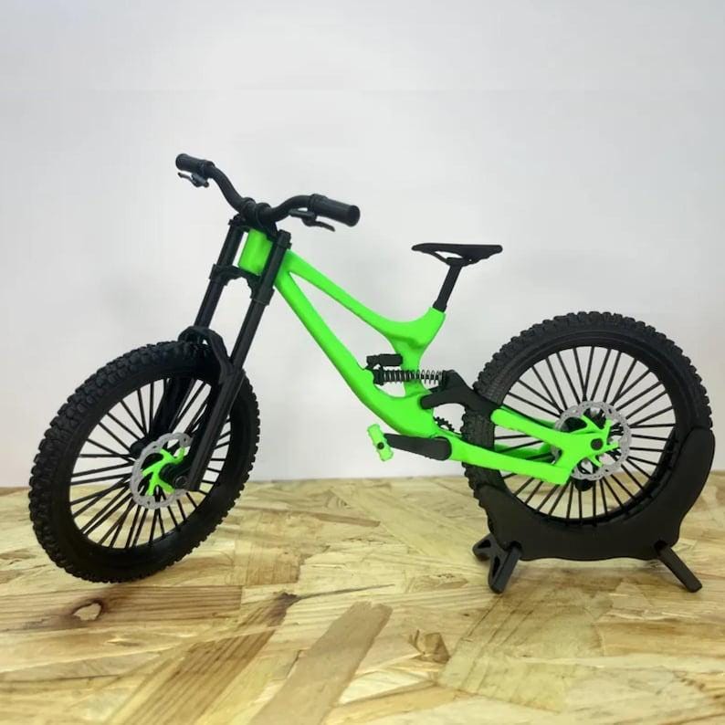 Pu&ograve; includere: Modello in miniatura di mountain bike con telaio verde neon e pneumatici neri. La bici presenta componenti dettagliati, tra cui sospensioni, freni a disco e sella nera. Il modello &egrave; esposto su una superficie in legno.
