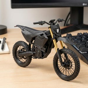 Puede incluir: Una motocicleta negra en miniatura con horquillas delanteras doradas. El modelo tiene un dise&ntilde;o detallado, que incluye neum&aacute;ticos, un asiento y un manillar. La motocicleta se exhibe sobre una superficie de madera cerca de un teclado y un monitor de computadora.