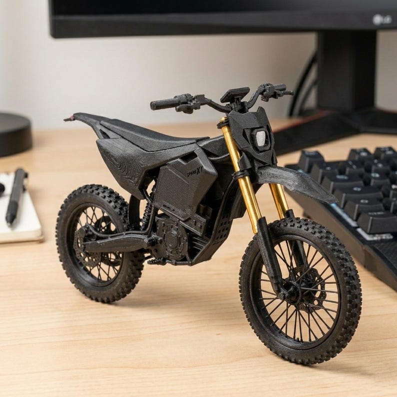 Puede incluir: Una motocicleta negra en miniatura con horquillas delanteras doradas. El modelo tiene un dise&ntilde;o detallado, que incluye neum&aacute;ticos, un asiento y un manillar. La motocicleta se exhibe sobre una superficie de madera cerca de un teclado y un monitor de computadora.