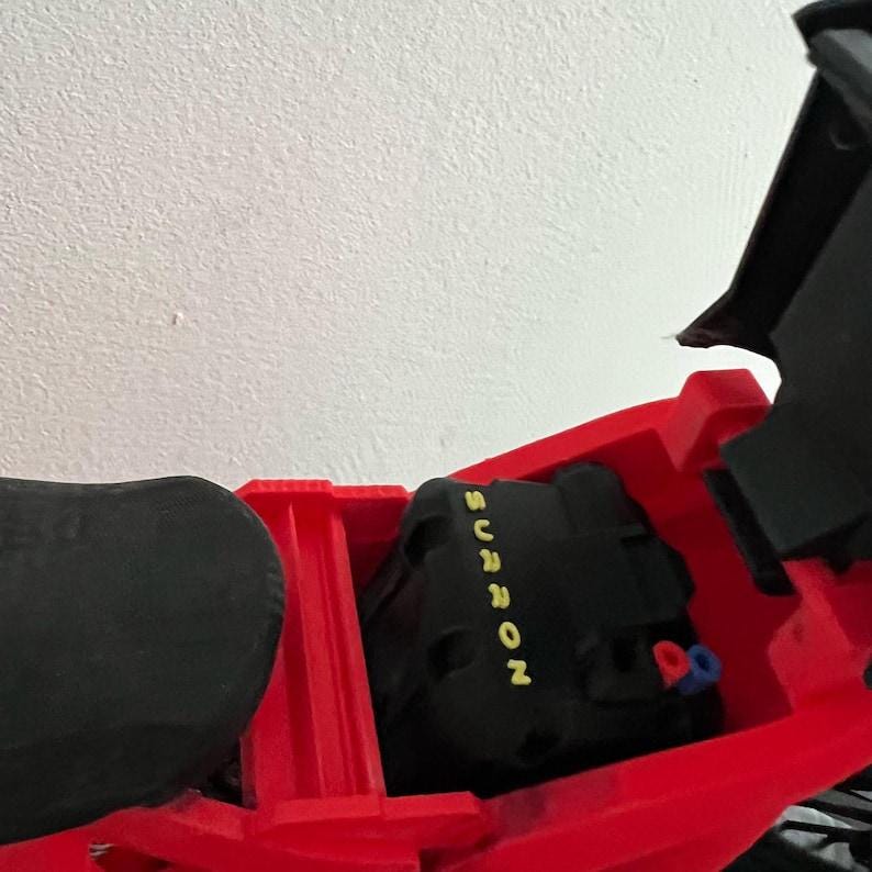 K&ouml;nnte beinhalten: Ein roter Elektro-Motorradrahmen mit einer schwarzen Komponente mit der gelben Aufschrift "SURRON". Das Motorrad hat einen schwarzen Sitz und eine schwarze Komponente im Rahmen. Der Hintergrund ist eine wei&szlig;e Wand.