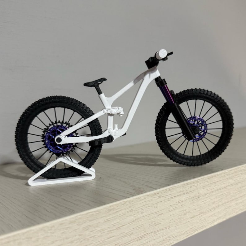 May include: A white and black miniature mountain bike with purple accents on the wheels and suspension. The bike has a detailed frame and tires, and is displayed on a white stand. The bike is a model or toy.