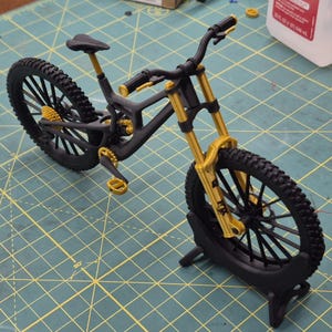 Könnte beinhalten: Ein detailliertes Modell eines Mountainbikes in Schwarz und Gold. Das Fahrrad hat schwarze Reifen, Rahmen und Lenker mit goldenen Details an Federung, Gängen und Pedalen. Es wird auf einem schwarzen Ständer präsentiert.
