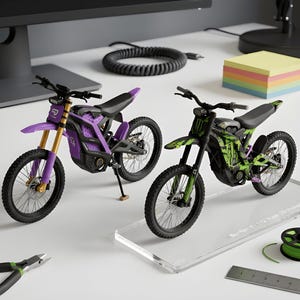 以下が含まれることがあります： 紫と緑のアクセントが入った黒の2台の電動ダートバイクのミニチュアモデル。詳細なハンドルバー、ブロックタイヤ、サスペンションフォークが特徴です。黒いモデルは、Sur-Ron X 1:12 Scaleと記された透明なアクリルベースの上に設置されています。