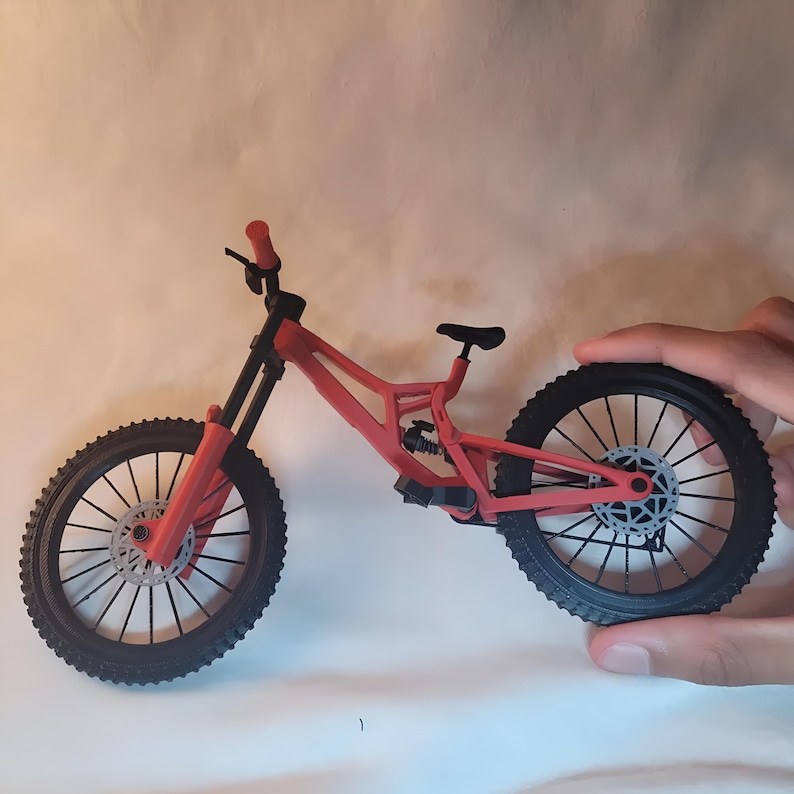 K&ouml;nnte beinhalten: Ein rotes und schwarzes Modell eines Mountainbikes mit grobstolligen Reifen und Scheibenbremsen. Das Fahrrad hat eine Federung und einen schwarzen Sattel. Der Lenker ist rot, und der Rahmen ist leuchtend rot. Es ist ein Miniatur-Nachbau.