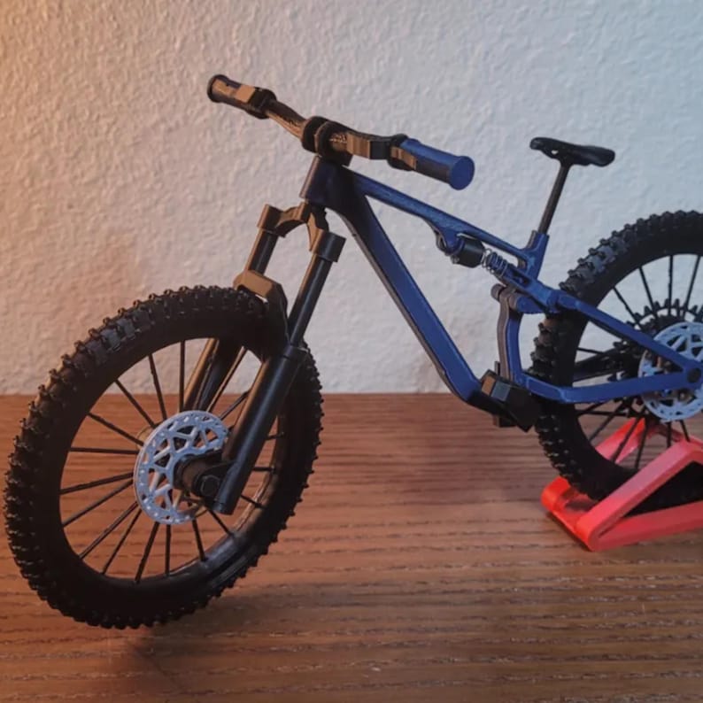 May include: A miniature blue mountain bike with black tires and a black seat. The bike has a detailed frame and is displayed on a red stand. The wheels have silver disc brakes.