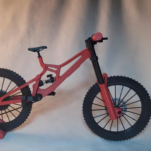 K&ouml;nnte beinhalten: Ein rotes Mountainbike-Modell mit schwarzen Reifen und einem schwarzen Sattel. Das Fahrrad verf&uuml;gt &uuml;ber ein Federungssystem und Scheibenbremsen. Der Lenker hat rosa Griffe. Das Modell ist eine Miniatur.