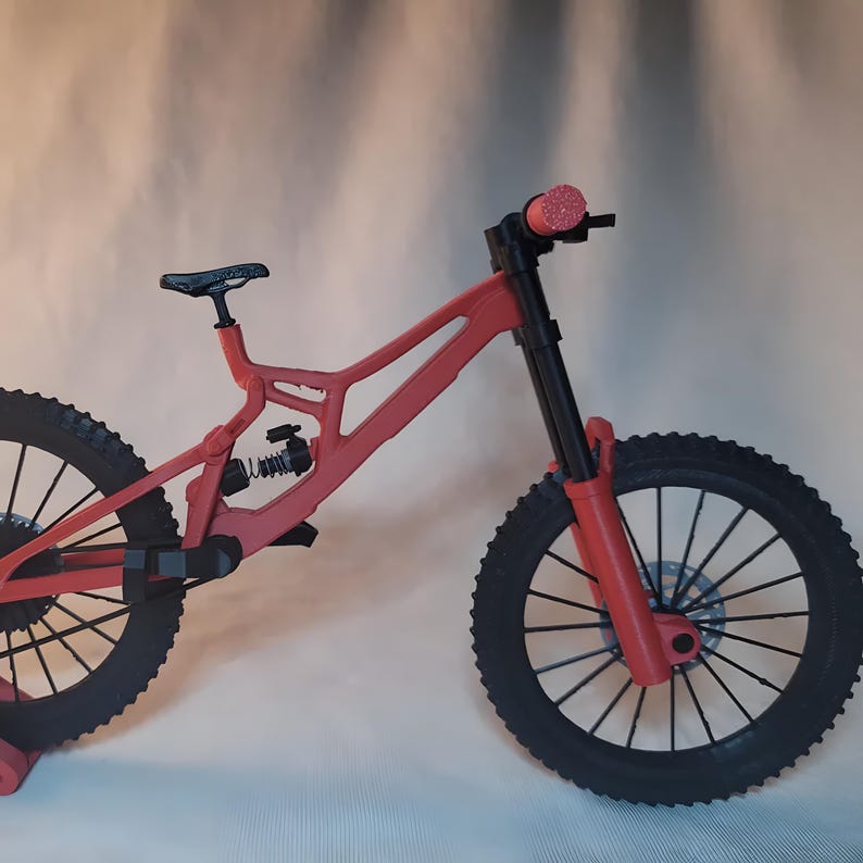 K&ouml;nnte beinhalten: Ein rotes Mountainbike-Modell mit schwarzen Reifen und einem schwarzen Sattel. Das Fahrrad verf&uuml;gt &uuml;ber ein Federungssystem und Scheibenbremsen. Der Lenker hat rosa Griffe. Das Modell ist eine Miniatur.