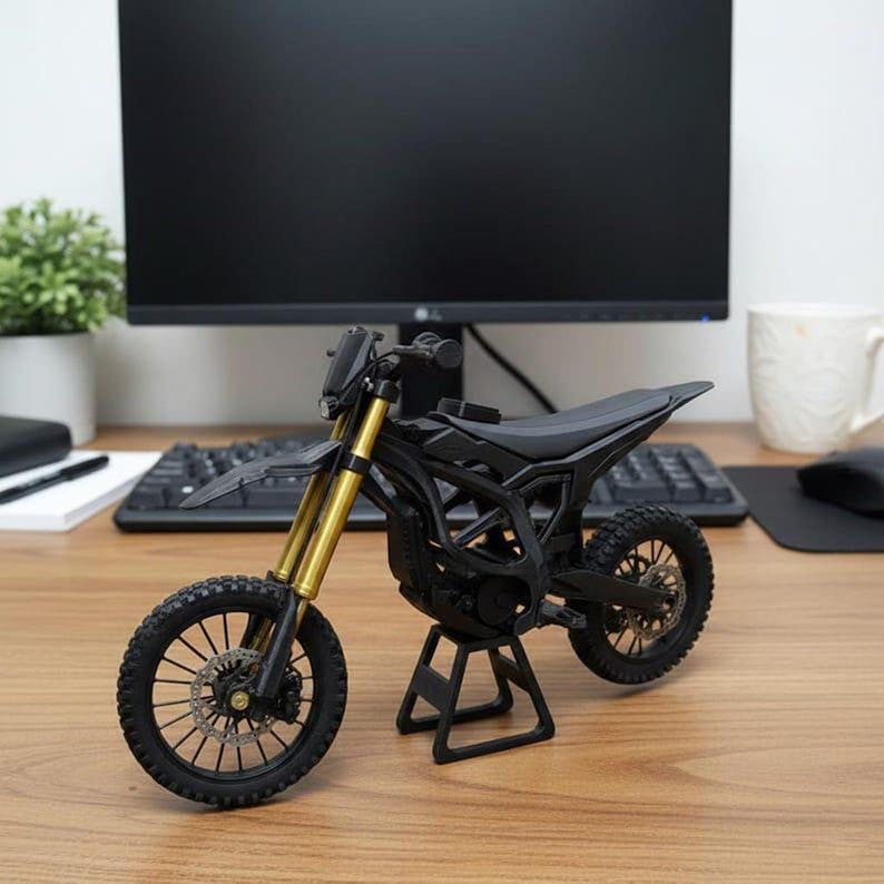 May include: A black and gold miniature motorcycle model. The model features a black frame, tires, and seat, with gold-colored front forks. The motorcycle is displayed on a black stand, showcasing its design and details.