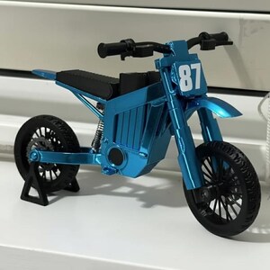 Puede incluir: Un modelo de motocicleta azul met&aacute;lico en miniatura con neum&aacute;ticos y manillar negros. El modelo presenta el n&uacute;mero "87" en la parte delantera y un asiento negro. La motocicleta se exhibe sobre una superficie blanca.