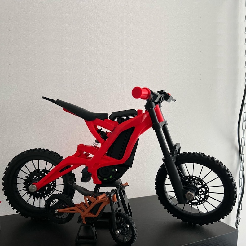 May include: A red and black model of an electric dirt bike with a smaller orange model in front. The larger bike has black tires, a black seat, and red frame. The handlebars and grips are red. The smaller bike is orange and black.