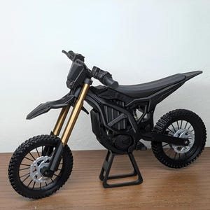 May include: A black and gold miniature motorcycle model with detailed tires, suspension, and frame. The model is displayed on a stand, showcasing its design. The motorcycle has a sleek, modern aesthetic.