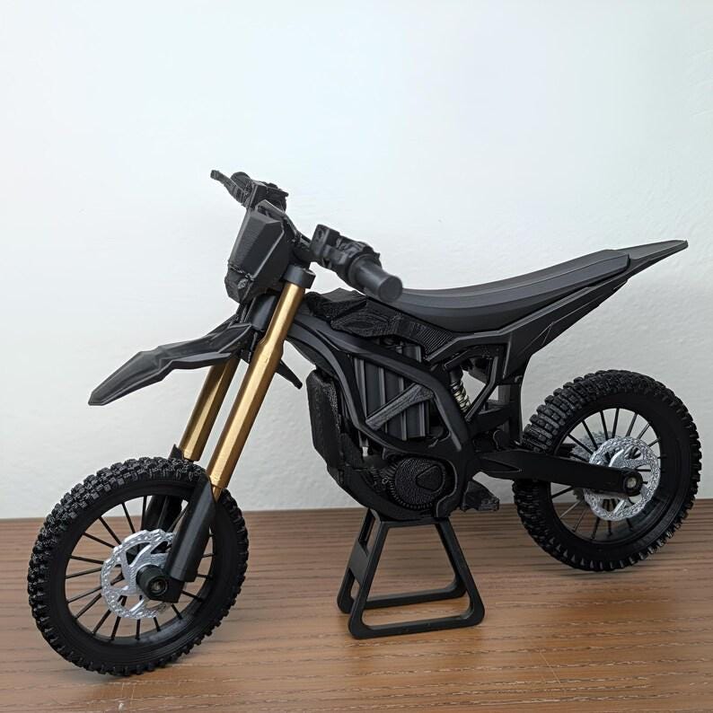 May include: A black and gold miniature motorcycle model with detailed tires, suspension, and frame. The model is displayed on a stand, showcasing its design. The motorcycle has a sleek, modern aesthetic.