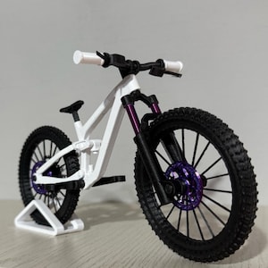 Könnte beinhalten: Weißes und schwarzes Mountainbike-Modell mit lila Akzenten an der Vorderradaufhängung und den Rädern. Die Reifen sind schwarz mit einem strukturierten Profil. Das Modell wird auf einem weißen Ständer präsentiert.