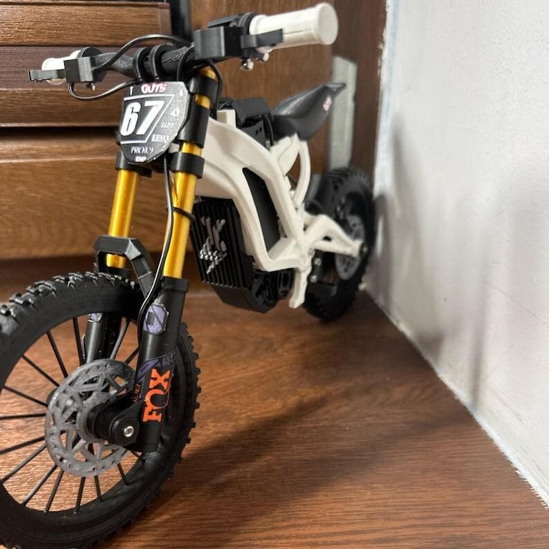 K&ouml;nnte beinhalten: Ein wei&szlig;es und schwarzes Spielzeug-Elektromotorrad mit goldenen Federgabeln und schwarzen Reifen. Das Vorderrad hat eine silberne Scheibenbremse. Die Nummer "67" ist auf einem kleinen Schild zu sehen. Die Marke "FOX" ist auf der Vorderradaufh&auml;ngung aufgedruckt.