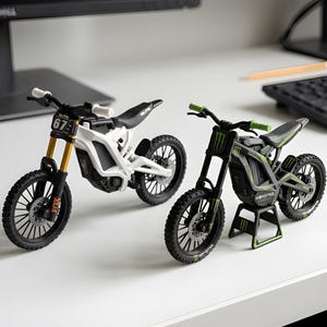 May include: Two miniature electric dirt bike models. The left model is white with gold front forks and the number 67 on the front plate. The right model is black with green accents, Monster Energy branding, and is mounted on a small black stand.