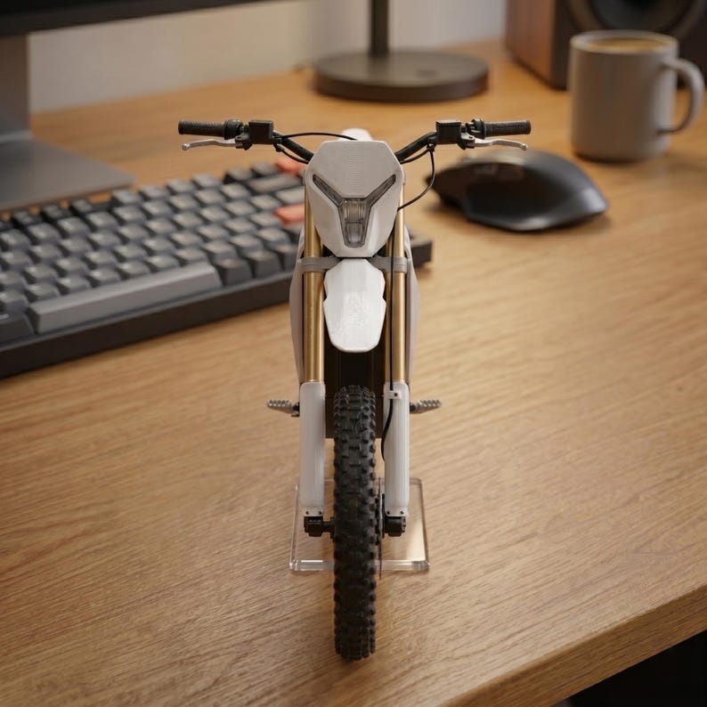 Op de afbeelding: Een miniatuurmodel van een witte en grijze motorfiets met goudkleurige veervorken. De motorfiets heeft een zwarte band en stuur. Het model staat op een transparante standaard op een houten bureau, met een toetsenbord en muis op de achtergrond.