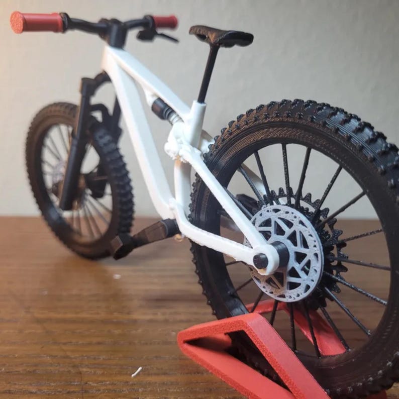 May include: A white and black mountain bike model with red handlebar grips. The bike has a black seat, tires, and suspension components. The rear wheel features a white disc brake and is supported by a red stand.