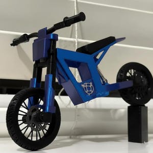 Puede incluir: Una motocicleta de juguete azul con neum&aacute;ticos y manillar negros. La motocicleta tiene un dise&ntilde;o de marco &uacute;nico y un asiento negro. Las ruedas son negras con patrones de radios detallados. La motocicleta se exhibe sobre un soporte negro.