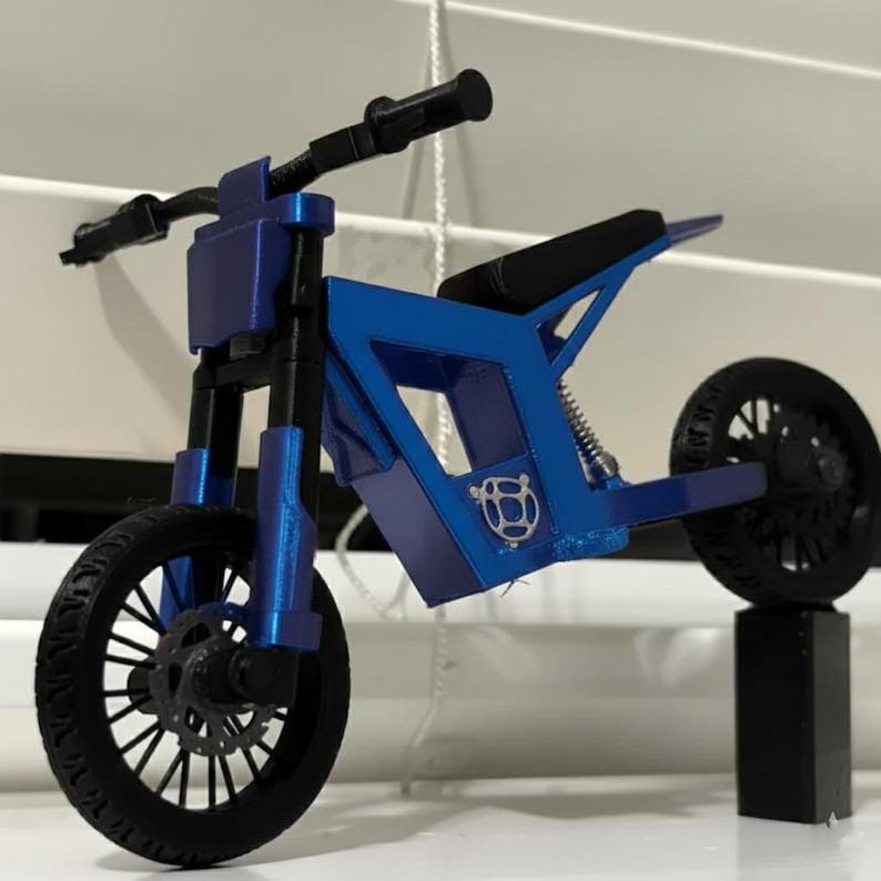 Puede incluir: Una motocicleta de juguete azul con neum&aacute;ticos y manillar negros. La motocicleta tiene un dise&ntilde;o de marco &uacute;nico y un asiento negro. Las ruedas son negras con patrones de radios detallados. La motocicleta se exhibe sobre un soporte negro.
