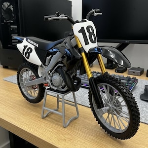 Peut inclure: Modèle de moto tout-terrain Honda noir et blanc avec le numéro 18 à l'avant et sur les panneaux latéraux. Le modèle a des fourches avant dorées et est exposé sur un petit support gris. Les pneus ont une bande de roulement à crampons.