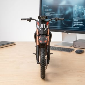 May include: A black and copper-colored toy motorcycle with the text "X1" on the front. The motorcycle has a black tire and is sitting on a wooden surface. The background includes a computer monitor, keyboard, and mouse.