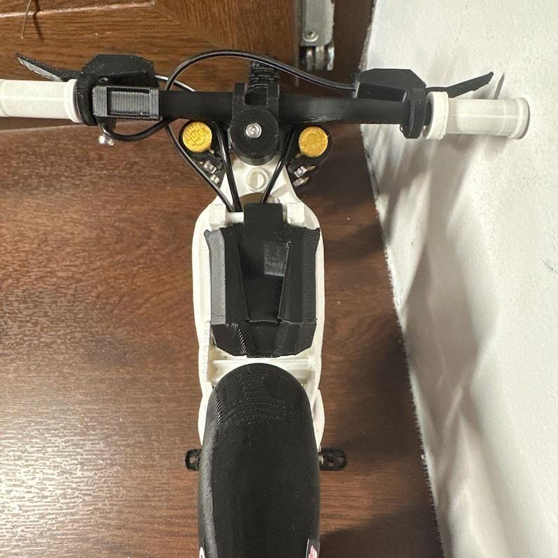 K&ouml;nnte beinhalten: Nahaufnahme eines wei&szlig;en und schwarzen Elektrorollers. Der Lenker ist schwarz mit wei&szlig;en Griffen. Der Roller hat gelbe Lichter und ein schwarzes Vorderrad. Der Roller steht auf einer Holzoberfl&auml;che.