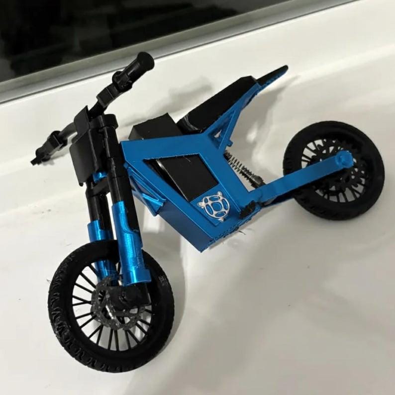 Puede incluir: Modelo de motocicleta en miniatura azul y negro con un dise&ntilde;o detallado. El modelo presenta un asiento, manillar y neum&aacute;ticos negros, con un marco y suspensi&oacute;n azules. Las ruedas tienen intrincados patrones de radios.