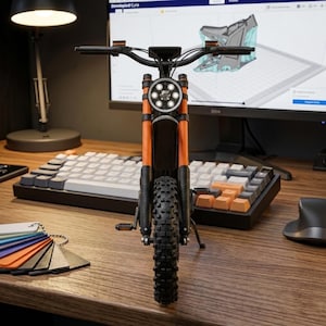 K&ouml;nnte beinhalten: Ein Miniaturmodell eines orangefarbenen und schwarzen Motorrads mit rundem Scheinwerfer, schwarzen Reifen und Lenker. Das Modell steht auf einem Holzschreibtisch mit Tastatur, einem Computermonitor mit 3D-Design und Farbmuster.