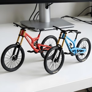 May include: Two miniature mountain bike models featuring detailed frames, black tires with spokes, and gold-colored front suspension forks. The red bike displays the Santa Cruz brand name on the frame, while the blue bike features the Evil brand name.