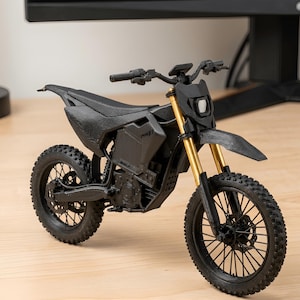 Puede incluir: Una miniatura de moto de cross negra con horquillas delanteras doradas. El modelo tiene un dise&ntilde;o detallado, que incluye neum&aacute;ticos, asiento, manillar y faro. La moto se exhibe sobre una superficie de madera clara.