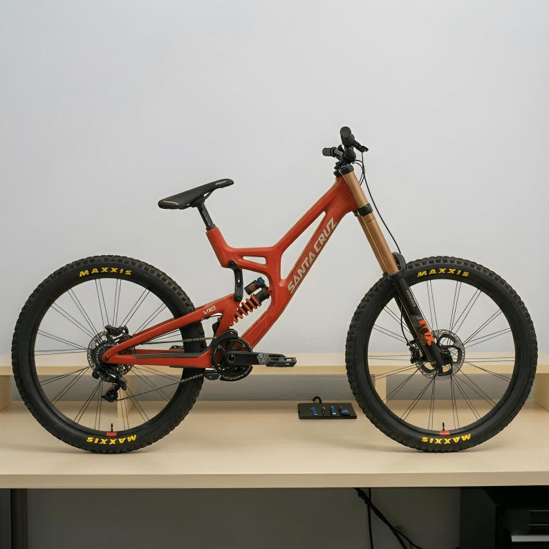 May include: A matte orange Santa Cruz V10 mountain bike featuring a full-suspension frame, black Maxxis tires with yellow lettering, a rear coil shock, front suspension forks, a black seat, and black pedals. The frame displays the brand name.