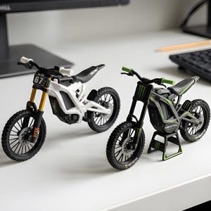 May include: Two miniature dirt bike models. One is white with gold front forks and the number 67 on the front plate. The other is black with green accents, Monster Energy branding, and is mounted on a small black and green display stand.