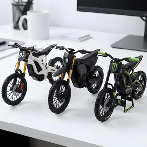 Può includere: Tre modelli in miniatura di moto da cross su una superficie bianca. La moto a sinistra è bianca con forcelle dorate e il numero 67. Quella centrale è nero opaco con forcelle dorate. Quella a destra è nera con accenti verdi e marchio Monster Energy.
