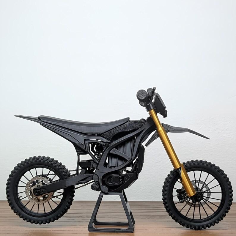 May include: A black and gold miniature dirt bike model. The model features a black frame, seat, and tires, with gold-colored front forks. The bike is displayed on a black stand, showcasing its intricate design and details.