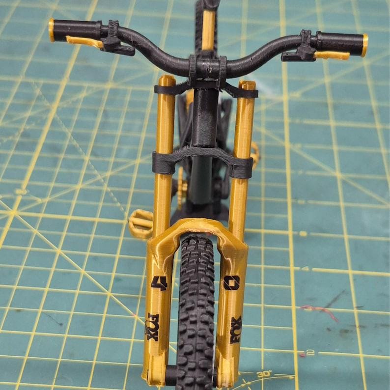 K&ouml;nnte beinhalten: Detaillierte Nahaufnahme eines Mountainbikes. Das Fahrrad hat einen schwarzen Rahmen, Lenker und Reifen mit goldfarbenen Federgabeln und Akzenten. Der Vorderreifen ist fokussiert und zeigt sein Profil. Das Fahrrad steht auf einer gr&uuml;nen Oberfl&auml;che.
