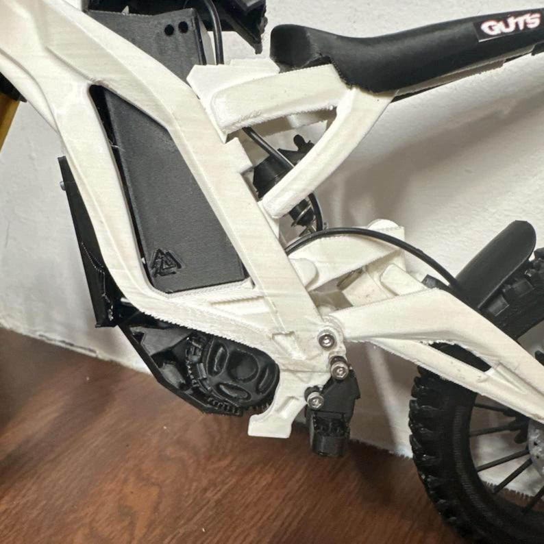 May include: Close-up of a white and black electric dirt bike. The bike has a black seat with the word "GUTS" printed on it. The frame is white, and the tires are black. The bike appears to be new.
