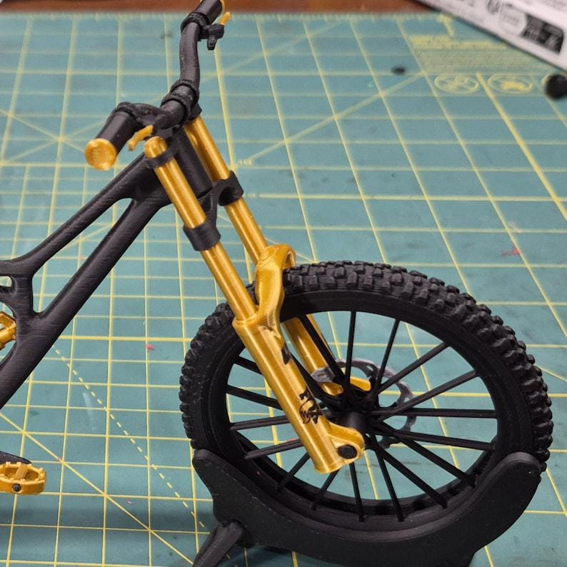 Puede incluir: Modelo en miniatura de bicicleta de monta&ntilde;a negra y dorada. La bicicleta presenta un cuadro negro, horquillas de suspensi&oacute;n doradas y un neum&aacute;tico negro con un dibujo detallado de la banda de rodadura. El manillar y los pedales tambi&eacute;n son dorados.