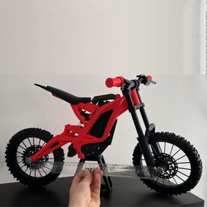 K&ouml;nnte beinhalten: Ein rotes und schwarzes Elektro-Dirtbike-Spielzeug mit schwarzen Reifen und einem schwarzen Sitz. Das Fahrrad hat einen roten Rahmen, Lenker und Akzente. Der Vorderradreifen hat einen Durchmesser von ca. 25 cm.