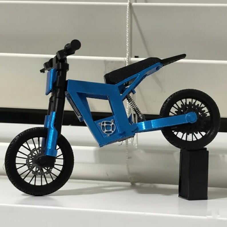 Puede incluir: Una peque&ntilde;a motocicleta de juguete azul con ruedas negras y un asiento negro. La motocicleta tiene un dise&ntilde;o de marco &uacute;nico y est&aacute; soportada por un soporte negro. Las ruedas tienen un patr&oacute;n de radios detallado.