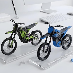 Puede incluir: Dos modelos de motos eléctricas de cross impresos en 3D sobre soportes transparentes. Una es verde y negra con el texto 3D Printed: Sur-Ron X - Green/Black. La otra es azul y negra con el texto 3D Printed: Electric Dirt Bike - Blue/Black.