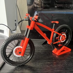 Peut inclure: Modèle réduit de vélo de montagne rouge et noir. Le vélo présente un cadre, un guidon et une selle rouges, avec des pneus et des roues noirs. Le modèle est exposé sur une surface noire.