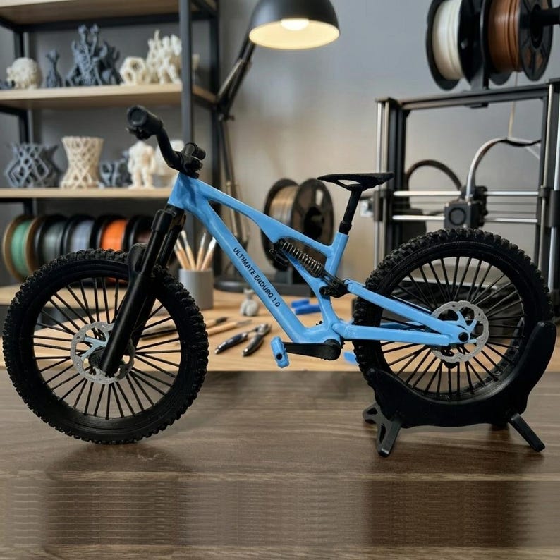 Puede incluir: Una bicicleta de monta&ntilde;a en miniatura de color azul claro con neum&aacute;ticos y componentes negros. La bicicleta tiene el texto "ULTIMATE ENDURO 1.0" en el cuadro. Se exhibe sobre una superficie de madera con un soporte negro.