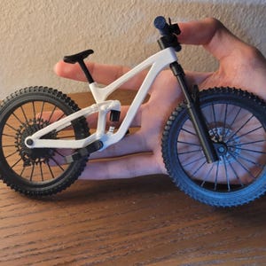 May include: A miniature white and black mountain bike with detailed tires, frame, and suspension. The bike is held in a hand, showcasing its small size and intricate design. The wheels have a diameter of approximately 3 inches.