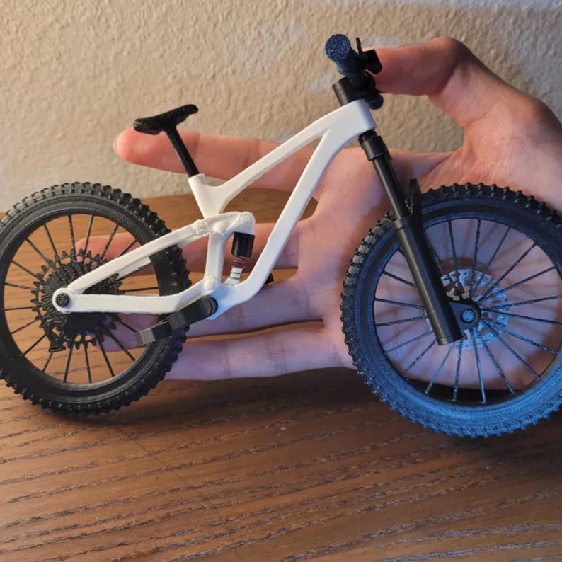 May include: A miniature white and black mountain bike with detailed tires, frame, and suspension. The bike is held in a hand, showcasing its small size and intricate design. The wheels have a diameter of approximately 3 inches.